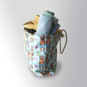 May include: A light blue canvas tote bag with a drawstring closure and a white rope handle. The bag is decorated with a repeating pattern of cartoon dogs wearing sunglasses and swimming trunks. The bag is filled with rolled up towels and a straw hat.