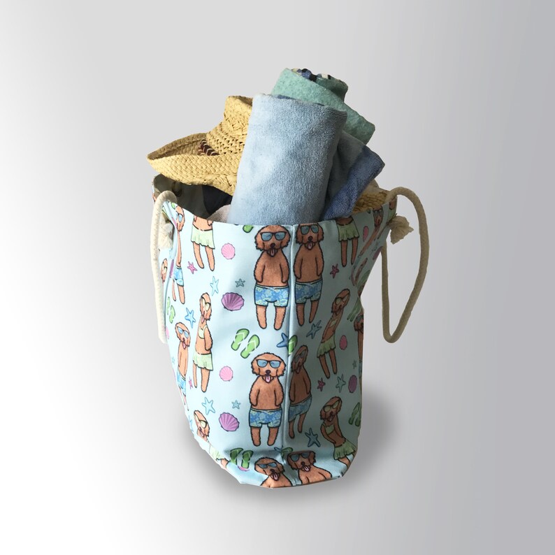 May include: A light blue canvas tote bag with a drawstring closure and a white rope handle. The bag is decorated with a repeating pattern of cartoon dogs wearing sunglasses and swimming trunks. The bag is filled with rolled up towels and a straw hat.