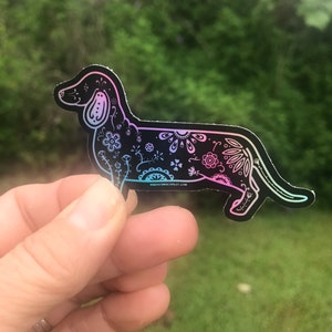 May include: A black and white dachshund silhouette sticker with a floral design. The sticker has a holographic finish that shifts between pink and blue.