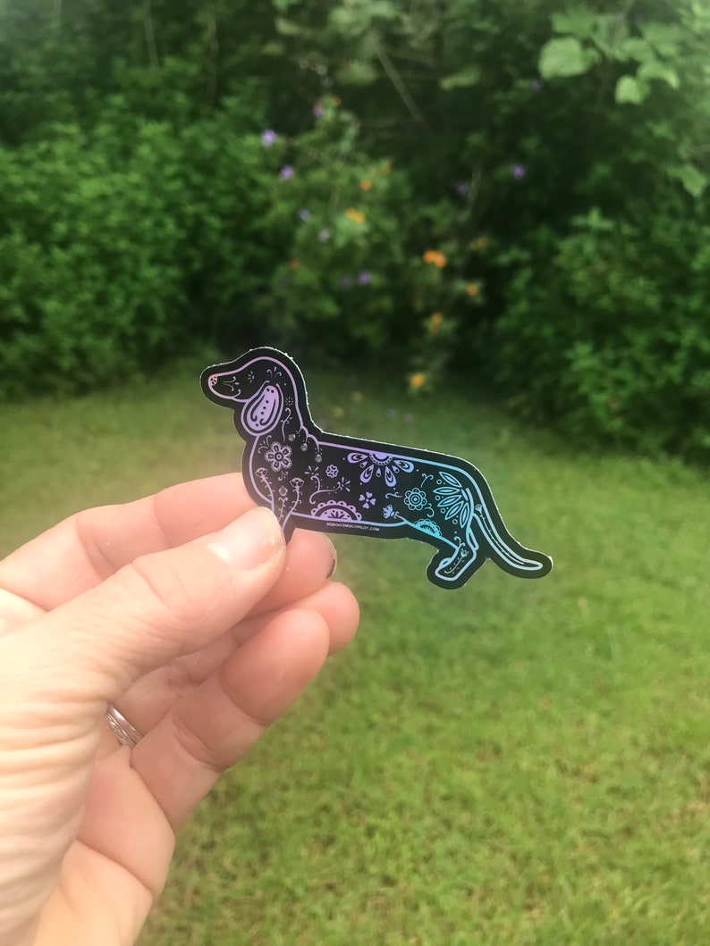 May include: A black and white dachshund sticker with a floral design. The sticker has a holographic finish that shifts from pink to blue.
