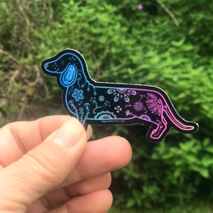 May include: A black vinyl sticker of a dachshund with a colorful floral pattern. The sticker has a gradient of blue and pink colors.
