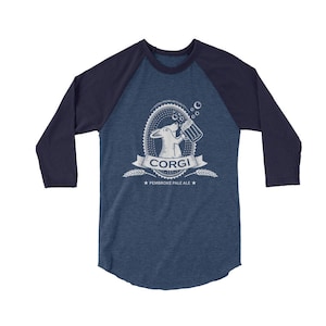 Puede incluir: Una camiseta de b&eacute;isbol azul y azul marino con un gr&aacute;fico blanco de un perro corgi bebiendo de una jarra de cerveza. El gr&aacute;fico incluye el texto "Corgi" y "Pembroke Pale Ale".