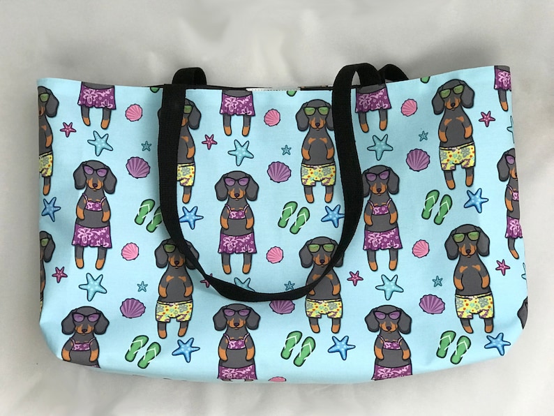 May include: A light blue tote bag featuring a repeating pattern of black and brown dachshunds wearing sunglasses and swimwear. The dachshunds are surrounded by seashells, starfish, and flip-flops.