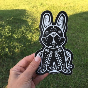 May include: Black and white sticker of a French bulldog with a Day of the Dead inspired design. The dog is sitting with its legs crossed and has flowers and leaves on its body. The sticker has a website address at the bottom.