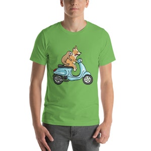 May include: A green t-shirt with a cartoon image of a corgi driving a blue scooter with a sloth riding on its back.
