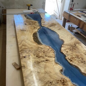 May include: A large wooden table with a blue epoxy river running through the center. The wood has a natural grain pattern and the epoxy is clear and shiny.