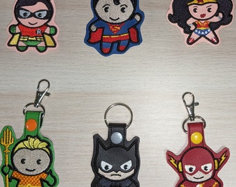 May include: Six embroidered superhero keychains with metal rings and clips. The keychains feature Superman, Wonder Woman, Robin, Aquaman, Batman, and The Flash. Each keychain is a different color and has a different design.