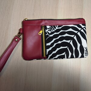 May include: A burgundy leather clutch with a black and white leaf print fabric panel and a gold zipper. The clutch has a detachable burgundy leather strap.
