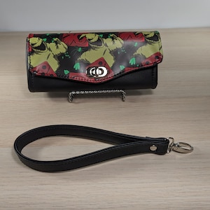 May include: A black wallet with a green, red, and black print of a cartoon character. The wallet has a black strap with a silver clasp.