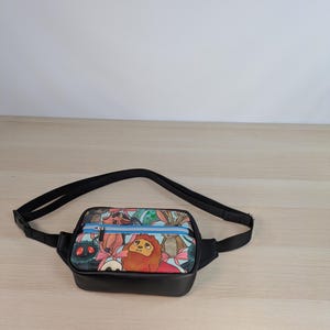 May include: A black fanny pack featuring a colorful cartoon print of various creatures, including a lion and a rabbit, with pink bows. The bag has a bright blue zipper across the front pocket and an adjustable black strap for waist or shoulder wear.