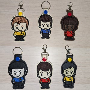 May include: Six embroidered keychains featuring cartoon characters from Star Trek. The characters are wearing different colored uniforms and have different colored hair. The keychains are attached to silver key rings.