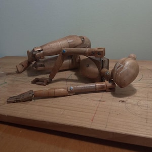 May include: Two wooden artist mannequins in a relaxed pose on a wooden surface. The mannequins are light brown with visible joints and screws. One mannequin's head has white markings. The background is a light blue-green.