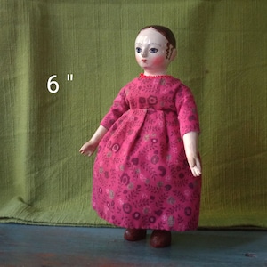 May include: A 6-inch tall wooden doll with painted features wearing a pink dress with a floral pattern. The doll has a red beaded necklace.