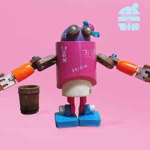 May include: A pink and wooden robot toy with blue and orange accents. The robot has a round body with the word "FRIEND" written on it. The robot has two arms and two legs. The robot is holding a small wooden bucket in its left hand.
