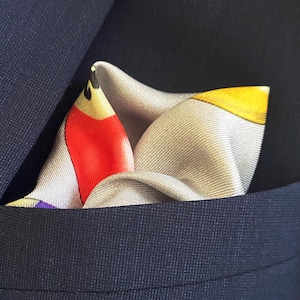 May include: A white pocket square with a colorful abstract design featuring red, yellow, and black accents. The pocket square is folded and placed in a suit jacket pocket.