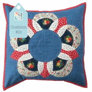 May include: Square blue cushion with a floral pattern. The cushion has a central blue circle with a scalloped edge, encircled by a flower-like design of red and white segments. The edges are trimmed in red, and a tag reads "Cushion Kit."