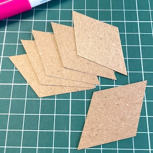 May include: Several diamond-shaped pieces of brown cardboard are arranged on a green grid surface. A pink and white marker is visible in the upper left corner. The cardboard pieces are likely for crafting or design projects.