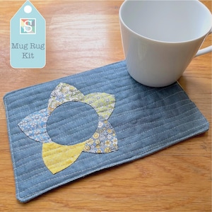 May include: A denim blue quilted mug rug with a floral applique. The applique features blue, yellow, and green floral patterns. A white ceramic mug sits on the rug. The tag reads "Mug Rug Kit".