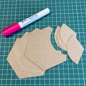 May include: A craft project setup featuring a pink and white glue pen and several brown paper cutouts in various shapes. The cutouts are arranged on a green grid cutting mat, suggesting a crafting or DIY project.
