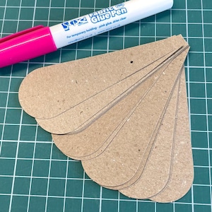 May include: A pink and white Stix2 Fabric Glue Pen lies near several brown, petal-shaped cardboard cutouts. The pen's text reads "For temporary holding - pink glue, dries clear." The cutouts are arranged on a green grid.