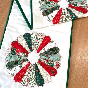 May include: White table runner with a green border and two decorative floral appliqués. The appliqués feature red, green, and white patterned fabric in a fan-like design, with a white centre and red stitching.