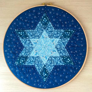 Puede incluir: Una obra de arte textil artesanal con un diseño de estrella de seis puntas. La estrella está compuesta por varias telas azules estampadas, cosidas sobre un fondo azul oscuro. La pieza está montada en un bastidor de bordado de madera, con detalles de estrellas blancas.