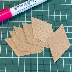 May include: A collection of diamond-shaped cardboard cutouts in various sizes, arranged on a green grid surface. A pink and white fabric glue pen is positioned at the top, with text that reads "Stixe Fabric Glue Pen".