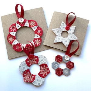Könnte beinhalten: Eine Sammlung handgefertigter Weihnachtsschmuckstücke. Die Ornamente umfassen einen Kranz, einen Stern und zwei blütenförmige Designs. Jedes Ornament besteht aus rotem, weißem und grauem Stoff mit Schneeflockenmustern und hat ein rotes Band zum Aufhängen.