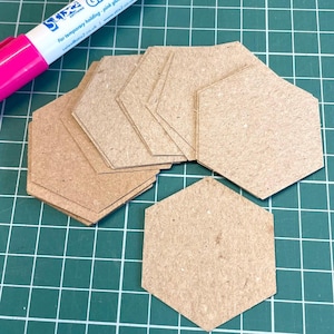 May include: A collection of hexagon-shaped cardboard pieces in various sizes. A pink marker with white text is in the upper left corner. The text on the marker reads "For temporary holding - pink glue". The pieces are on a green grid.