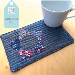 May include: A denim blue mug rug kit with a floral bow-tie design in red, white, and blue. The mug rug is rectangular and quilted, with the words "Mug Rug Kit" on a light blue tag. A white mug sits on the rug.
