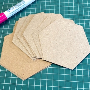 May include: A stack of hexagon-shaped, light brown cardboard tags. A pink and white glue pen is in the upper left corner. The tags are arranged on a green grid surface.