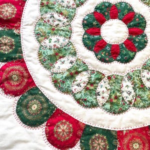 Op de afbeelding: Een close-up van een kerst-thema gewatteerde stof met een witte achtergrond. Het ontwerp heeft rode en groene cirkelvormige patronen met hulst- en bloemenprints, en een grote bloemvorm. De randen zijn omrand met rode stiksels.