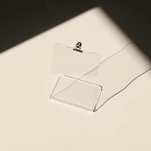 May include: A clear acrylic picture frame with a metal clip at the top. The frame is standing upright on a white surface.