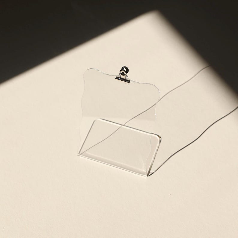 May include: A clear acrylic picture frame with a metal clip at the top. The frame is standing upright on a white surface.