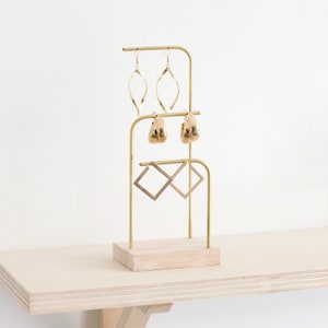 Op de afbeelding: Gouden metalen oorbellenstandaard met een houten basis. De standaard heeft drie niveaus voor oorbellen. Het bovenste niveau heeft twee paar gouden creolen, het middelste niveau heeft twee paar gouden hangende oorbellen en het onderste niveau heeft twee paar gouden vierkante oorbellen.