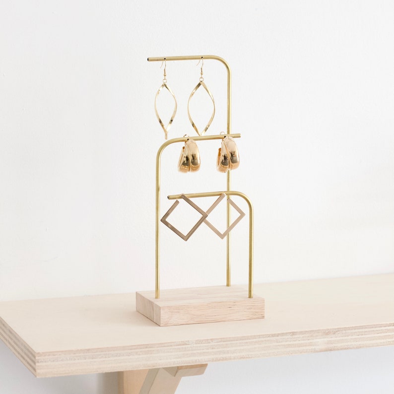 Op de afbeelding: Gouden metalen oorbellenstandaard met een houten basis. De standaard heeft drie niveaus voor oorbellen. Het bovenste niveau heeft twee paar gouden creolen, het middelste niveau heeft twee paar gouden hangende oorbellen en het onderste niveau heeft twee paar gouden vierkante oorbellen.