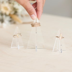 K&ouml;nnte beinhalten: Drei transparente Acryl-Ringst&auml;nder mit goldenen Ringen auf jedem. Die Ringst&auml;nder sind dreieckig.