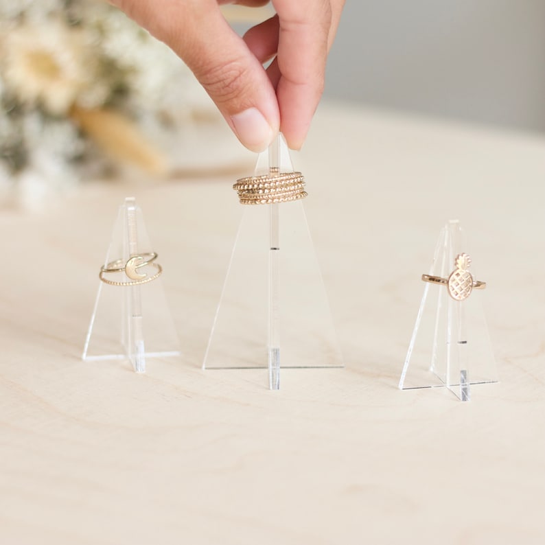K&ouml;nnte beinhalten: Drei transparente Acryl-Ringst&auml;nder mit goldenen Ringen auf jedem. Die Ringst&auml;nder sind dreieckig.