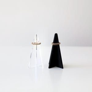 K&ouml;nnte beinhalten: Zwei Ringst&auml;nder, einer aus klarem Acryl und einer aus schwarzem Material, jeweils mit einem goldenen Ring obenauf.
