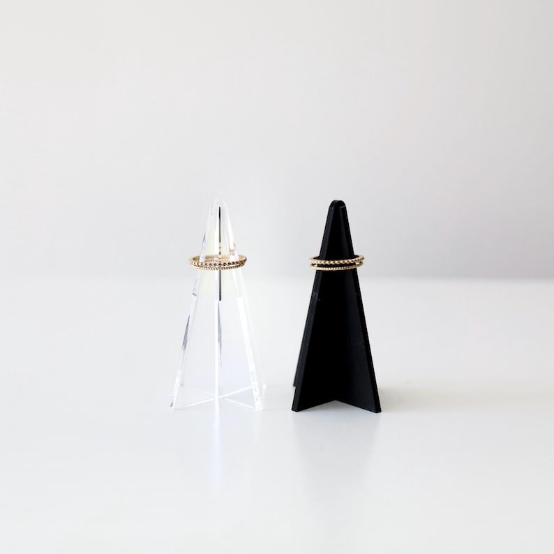 K&ouml;nnte beinhalten: Zwei Ringst&auml;nder, einer aus klarem Acryl und einer aus schwarzem Material, jeweils mit einem goldenen Ring obenauf.