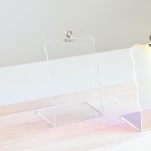 May include: Two clear acrylic display stands with a silver clip on top. The stand on the left has a wavy edge. The stand on the right has a iridescent finish.