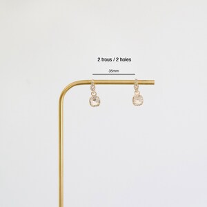 Op de afbeelding: Een paar gouden creolen met een vierkante kristallen hanger. De oorbellen zijn 35 mm lang en hebben twee gaten.