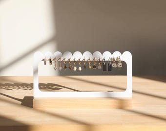 Puede incluir: Porta pendientes blanco con bordes festoneados, que muestra una variedad de pendientes de aro dorados con diferentes colgantes, incluyendo una hoja, una estrella, un círculo pequeño y un cristal.