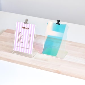 May include: Two clear acrylic sign holders with wavy edges. The left holder has a pink and white striped paper with a menu listing coffee and tea prices. The right holder is iridescent blue and green.