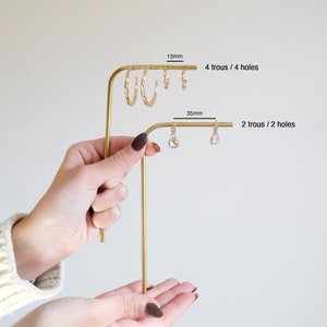 Op de afbeelding: Gouden metalen oorbellenstandaard met twee niveaus. De bovenste laag heeft vier gaten en is 3,8 cm lang. De onderste laag heeft twee gaten en is 3,5 cm lang.  "4 trous / 4 holes" en "2 trous / 2 holes" staan op de standaard.