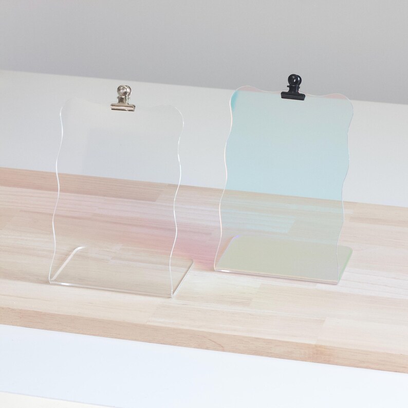 May include: Two clear acrylic display stands with wavy edges and clips on top. The stand on the left is pink and the stand on the right is blue.