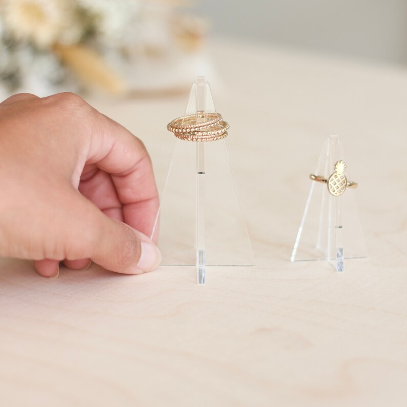 K&ouml;nnte beinhalten: Zwei transparente Acryl-Ringst&auml;nder. Ein St&auml;nder hat einen Stapel goldener Ringe, der andere hat einen einzelnen goldenen Ring mit einem Ananas-Design.