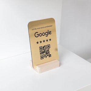 Puede incluir: Un letrero rectangular dorado con el logotipo de Google, cinco estrellas y un código QR. El letrero está montado sobre una pequeña base de madera clara. El texto "VOTRE AVIS NOUS INTERESSE!" está en la parte superior.