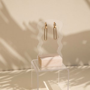 Op de afbeelding: Gouden creolen met een hangende rechthoekige vorm. De oorbellen worden gepresenteerd op een transparante acrylstandaard met een houten basis.