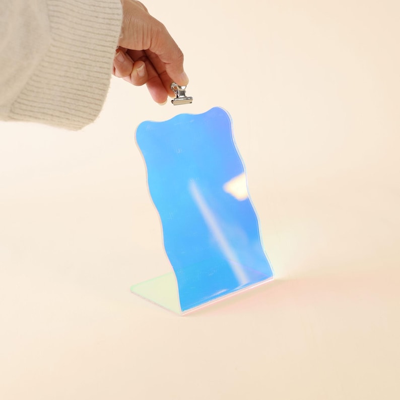 May include: A wavy iridescent blue mirror with a silver clip on top. The mirror is standing upright on a white surface.